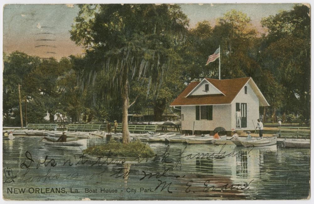 A circa 1905 postcard reproduced from a photograph showing the small boat house and lake in City Park; rows of row boats are tied up to the shore. In the foreground is a small island with a single tree dripping with Spanish moss.