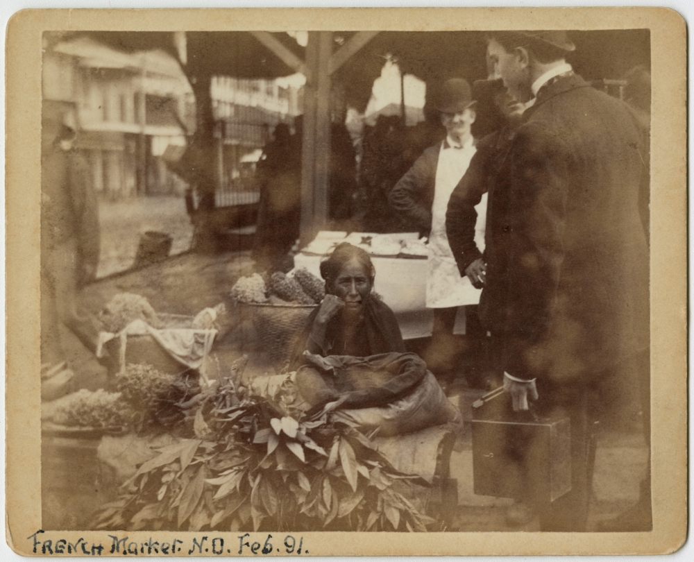 View of a vendor, a native American woman, seated at the French Market with baskets and bundles of various plant materials around her. Several men are also in view; one appears to also be a vendor and is wearing an apron. Buildings, probably along Decatur Street, are seen in the background.
