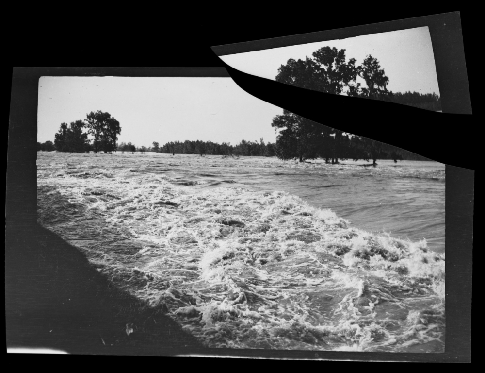 Black and white photo of flood waters at a shore line. The photo is ripped.