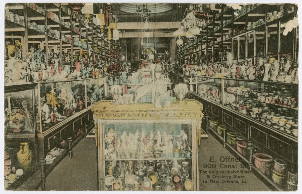 A circa 1910 postcard showing the interior of E. Offner, a glass and crockery store once located at 908 Canal Street. A glass display case stands in the center, and down both sides of the long store are more display cases. The store is jam-packed with merchandise not only in the cases but on shelves above and below as well as items on top of the glass cases. Decorative light fixtures hang from the ceiling.