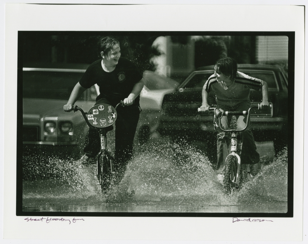 Two boys ride their bikes through a flooded street, smiling at each other
