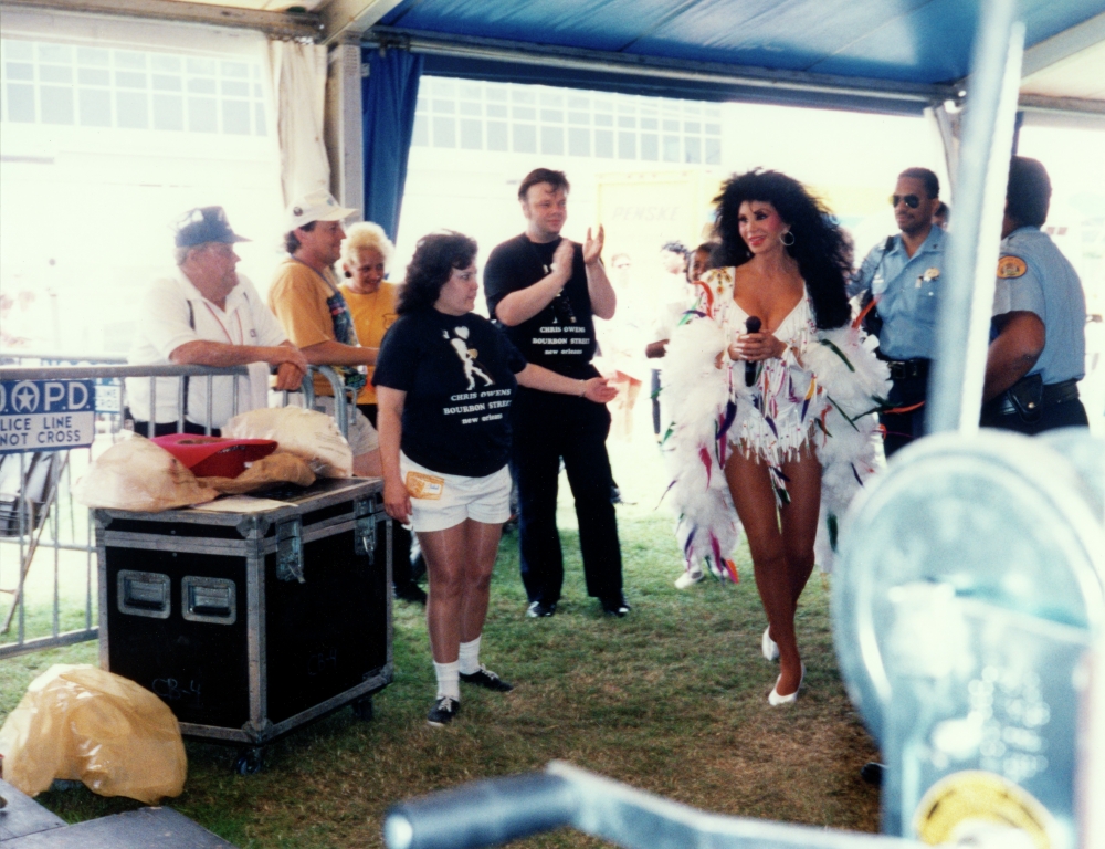Photograph of a dark-haired woman in an outdoor backstage area, holding a microphone and wearing a multicolored performance ensemble. 