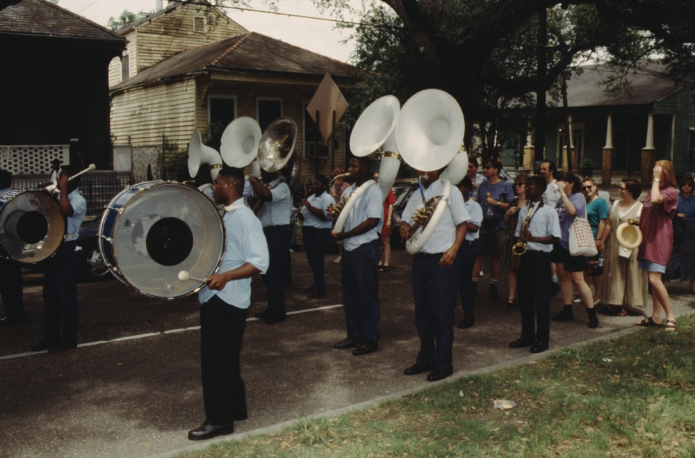 Color photograph of the drum and sousaphone sections of a marching band on a city street. A small crowd stands behind the band,