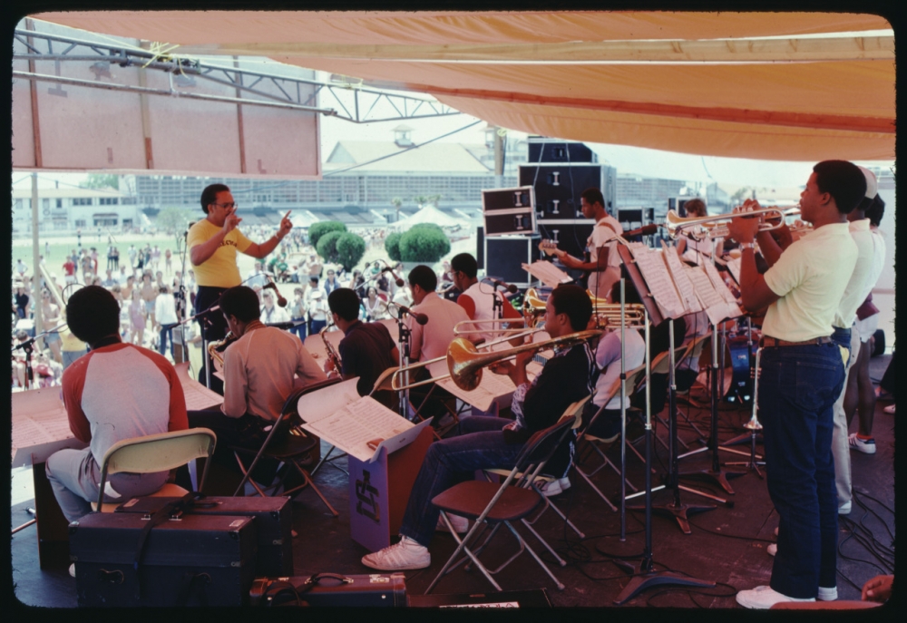 Three rows of band members face away from the camera, looking out over an audience from an outdoor stage.