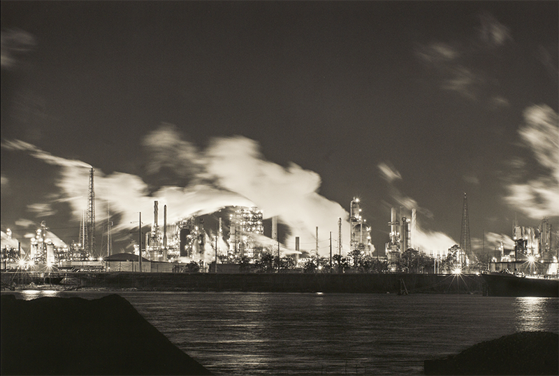 Image copyright of Richard Sexton; shared by THNOC. Black and white photograph of the petrochemical facilities in Norco, Louisiana. The facility's buildings, smoke stacks, and electrical grid shine lights onto the still water of the large, wide river.