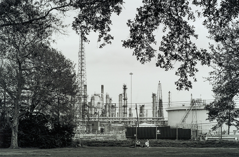 Image copyright of Richard Sexton, provided by THNOC. A black and white photograph show three young children playing in a greenspace alongside an oil refinery.