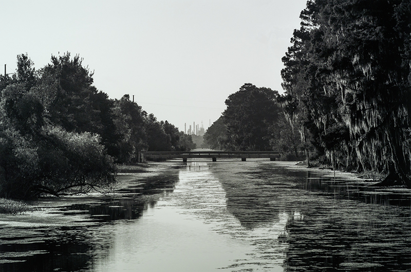 Image copyright of Richard Sexton; provided by THNOC. Black and white photograph showing a refinery in Norco, Louisiana. The image was taken from a drainage canal and there is calm water in the foreground.