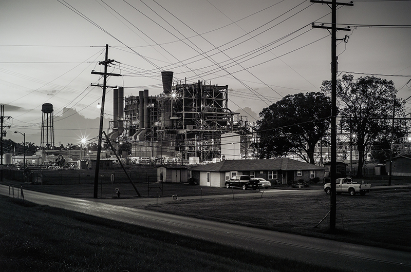 Image copyright of Richard Sexton; provided by The Historic New Orleans Collection. In this black and white photograph, a suburban residence is shown adjacent to a power plant.