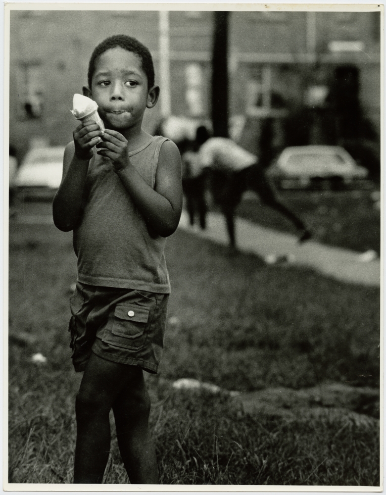 Boy eating a melting ice cream cone in a courtyard, Desire housing project buildings seen in background