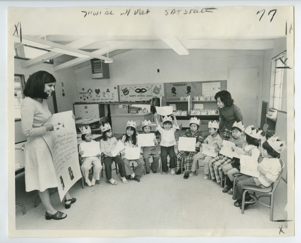 A black and white photo of a classroom. A female teacher stands at left holding a poster. A group of children are seated in chairs and wearing paper crowns. At left, behind the students, is another adult.