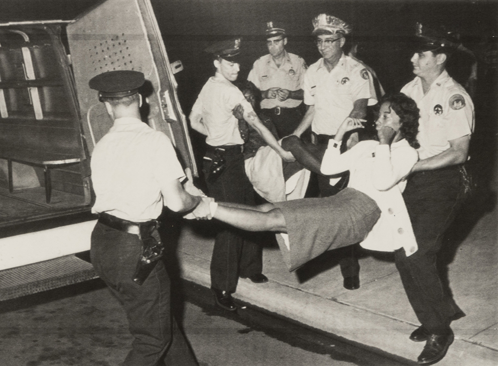 Black-and-white photograph of two young Black women being carried into a police van by five white police officers.