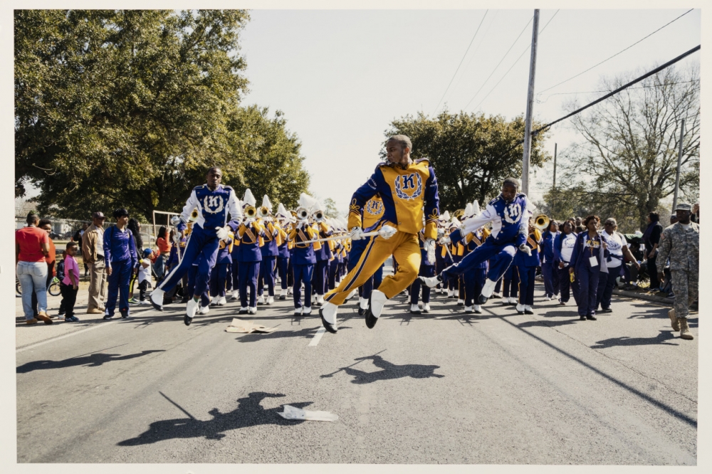 Color photograph of a marching band approaching the camera. In the foreground, three people are pictured in the air, mid-leap.