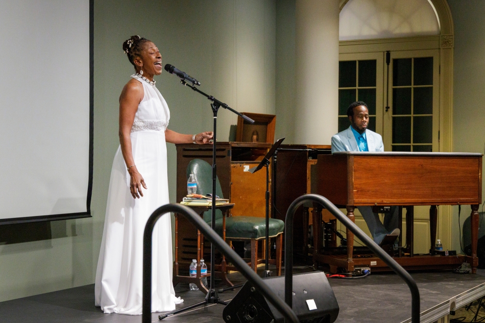 Vocalist Andaiye Alimayu stands at a microphone singing. To the right, Frederick “Jay” East accompanies her on piano.