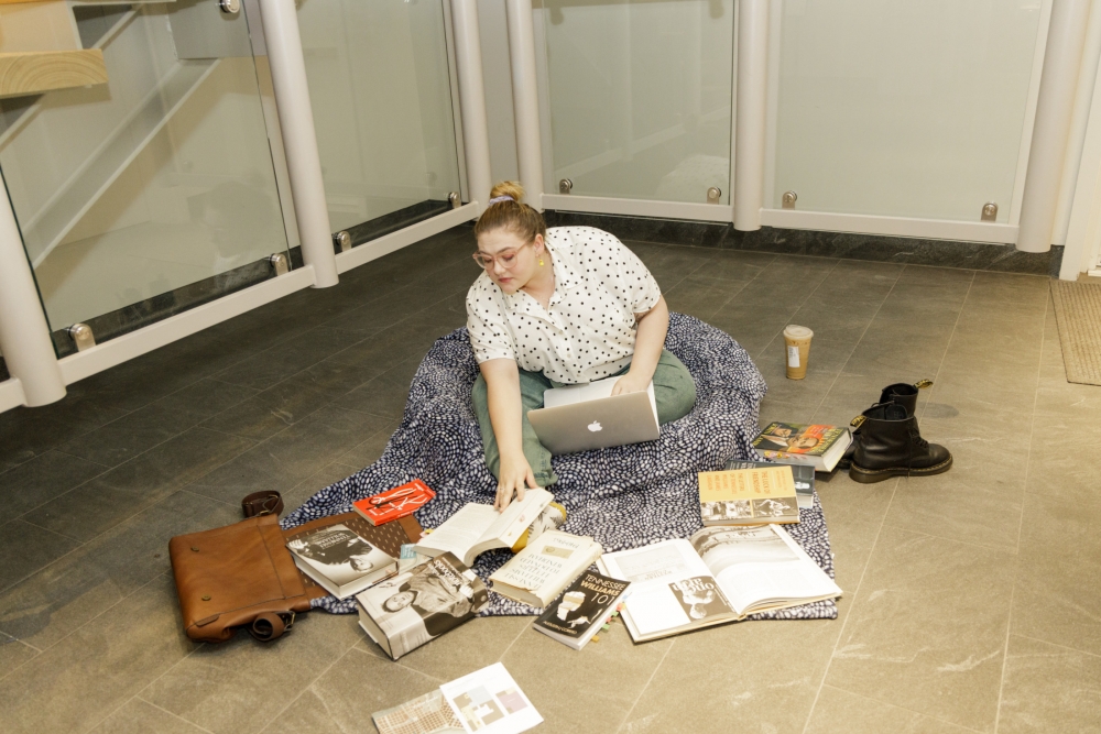Actress portraying "the scholar" sits on the floor, surrounded by books, laptop in her lap