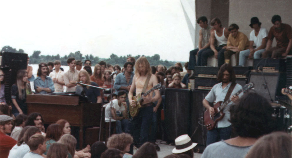 Color photograph of the Allman Brothers Band playing an outdoor concert on the riverfront, surrounded by spectators.