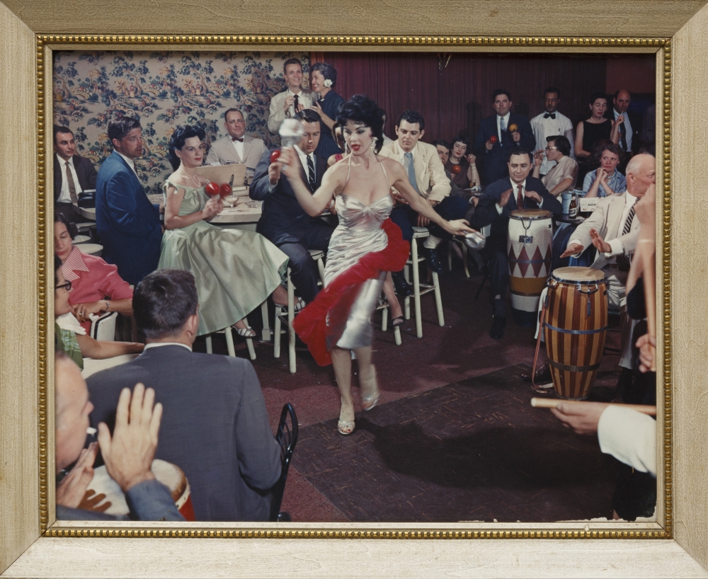 A framed color photograph of a dark-haired woman who is wearing a red and silver dress, dancing in a crowd.