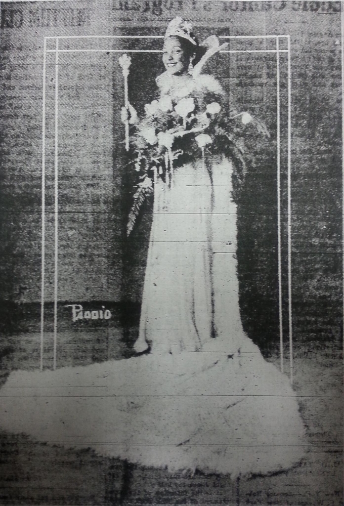 A black-and-white, three-quarters portrait of woman wearing a large, jeweled crown and a ball gown with a long train spread out in front of her. She holds a scepter in her right hand and cradles a bouquet of flowers and greenery in her left arm.
