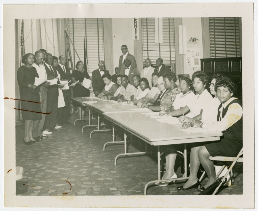 A group of Black women and men look at the camera. Some are seated at a long table. Others stand in at the far end of the table. Many are holding papers and pencils.