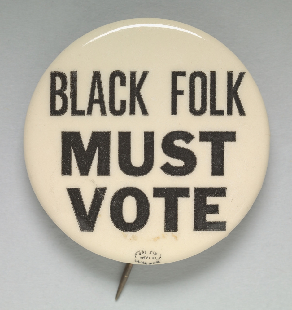 A white button with black texts that reads "BLACK FOLK MUST VOTE."
