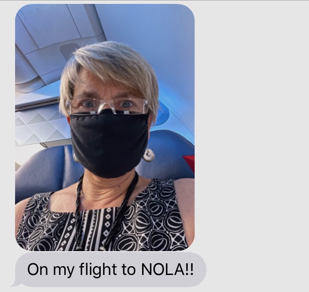 Screenshot of a text message consisting of a selfie of a woman wearing a mask over her nose and mouth, in a seat on a plane. Under the photo is the message "On my flight to NOLA!!"