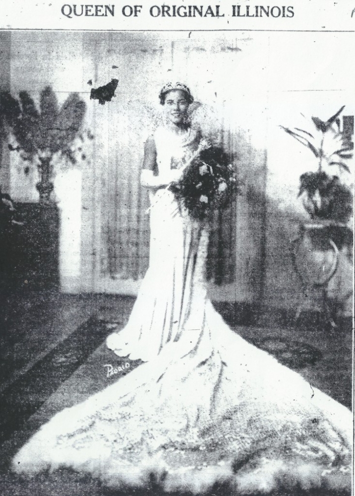 A black-and-white photo of young woman with a tiara on her head. She wears a white ball gown with a long train spread out on the floor in front of her. She holds a scepter in one hand and a large bouquet in the other.