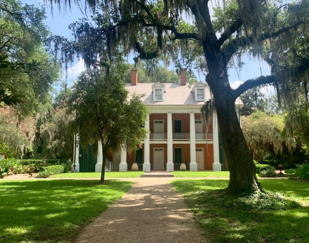 View of a red brick plantation house with white columns. Moss-covered trees stand in the foreground.