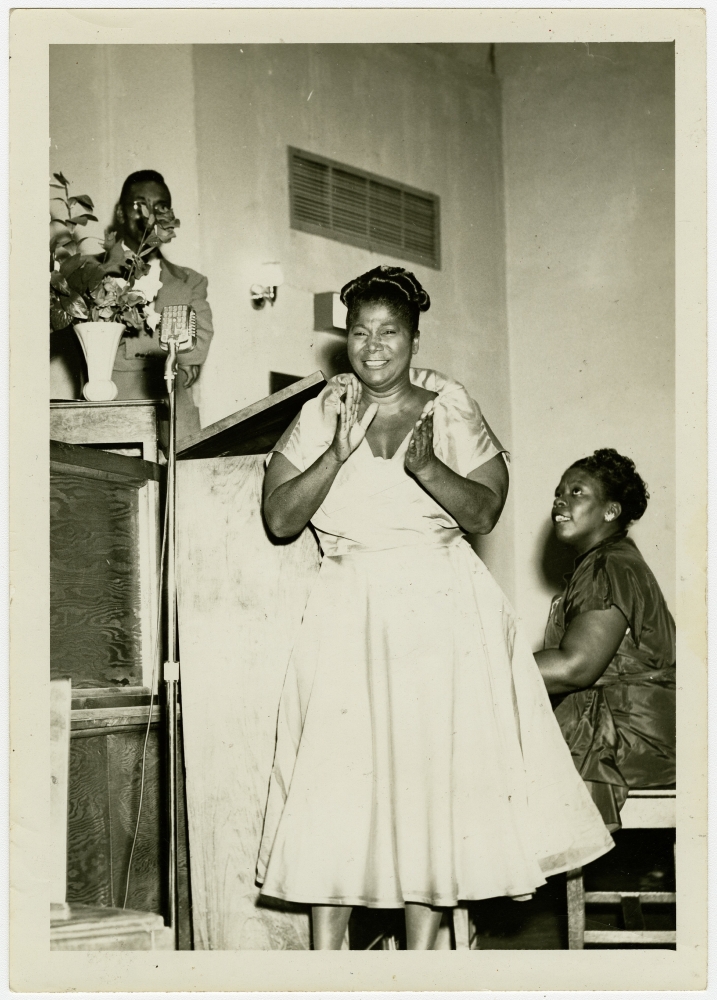 A black and white photo of Mahalia Jackson singing. Behind her, a woman plays paino.