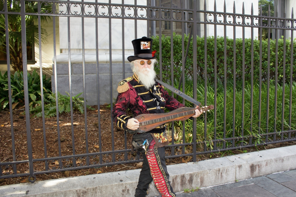 Full-length portrait of Amzie Adams standing on sidewalk, wearing black top hat, reflective sunglasses, marching band jacket, and pants, holding dulcimer