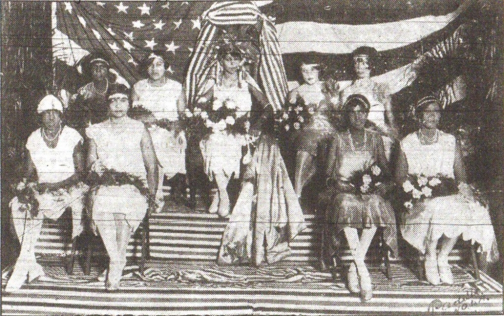 Nine young women are seated in two rows, with the queen in the center of the back row. They wear elegant, tea-length dresses, and each has a bouquet lying across her lap. A large US flag serves as a backdrop.