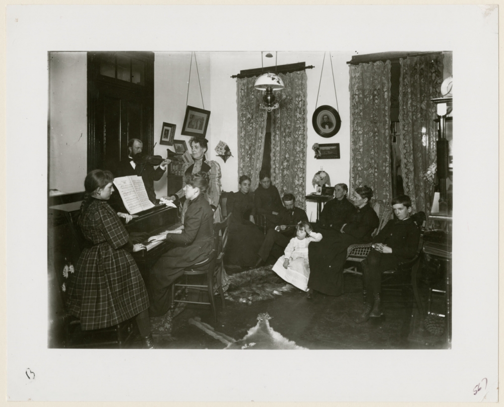  Photograph of a family in a parlor with a woman playing the piano, a man playing a violin, and a woman singing.