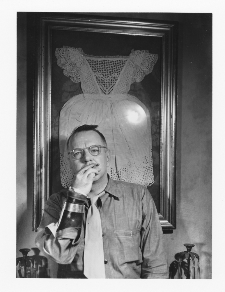 Black and white photograph of a man from the waist up. He is in front of a framed dress and is smoking a cigarette.